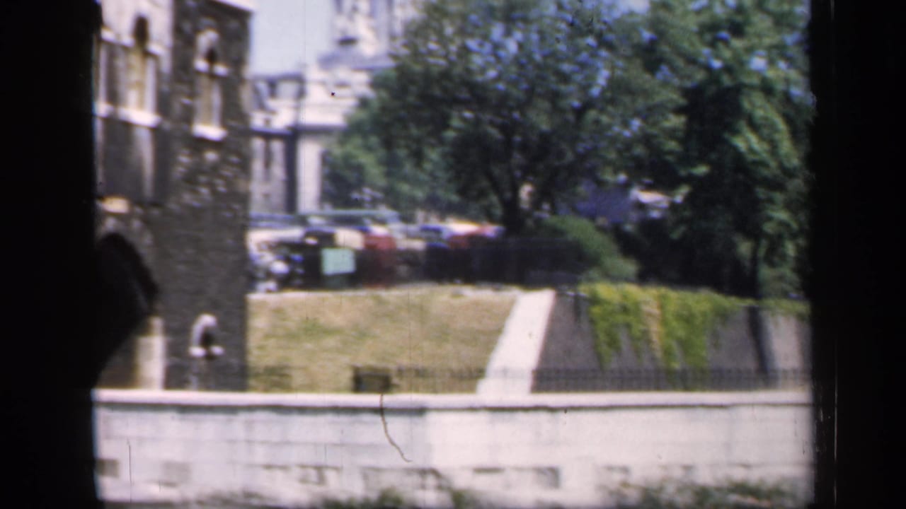 Color home-movie frame of an English historic building edge with grounds, trees, a path, and distant parked cars.