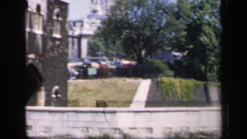 Color home-movie frame of an English historic building edge with grounds, trees, a path, and distant parked cars.