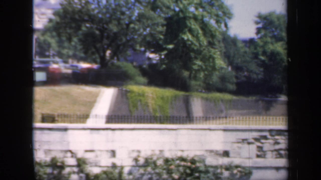 Park lawn, trees, pale retaining wall, and a path beside water or a lower embankment.