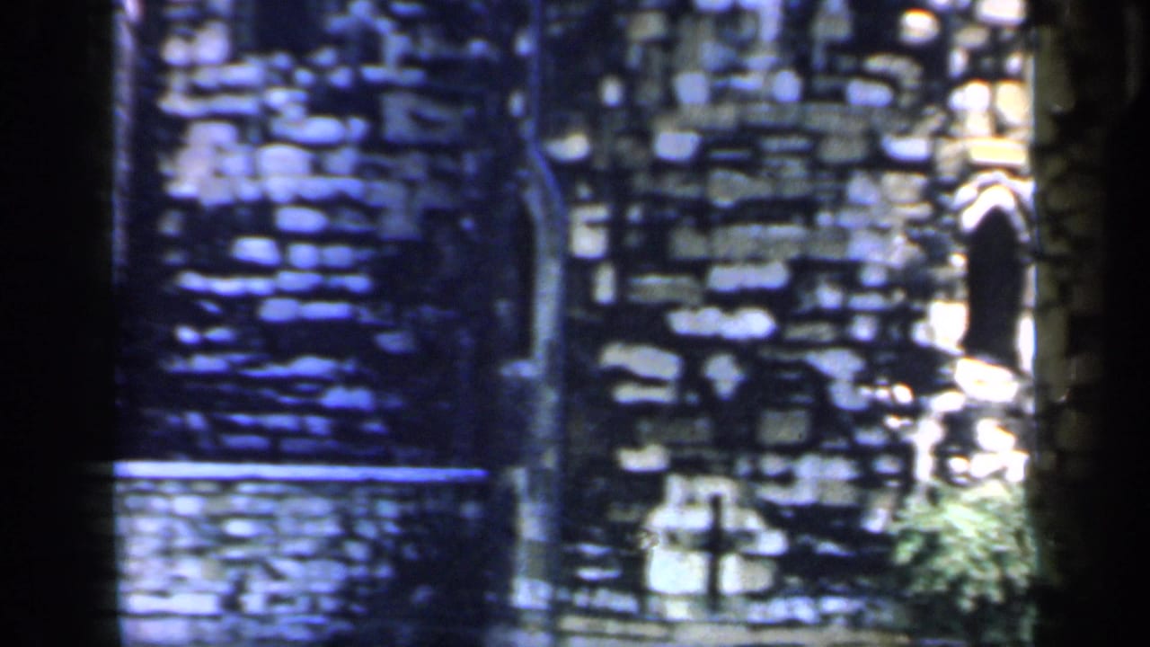 1949 8mm frame of dark Tower of London stonework with a sunlit lower wall, greenery, and a bright opening at the right edge.