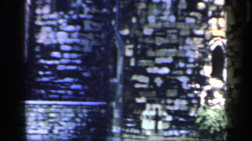 1949 8mm frame of dark Tower of London stonework with a sunlit lower wall, greenery, and a bright opening at the right edge.