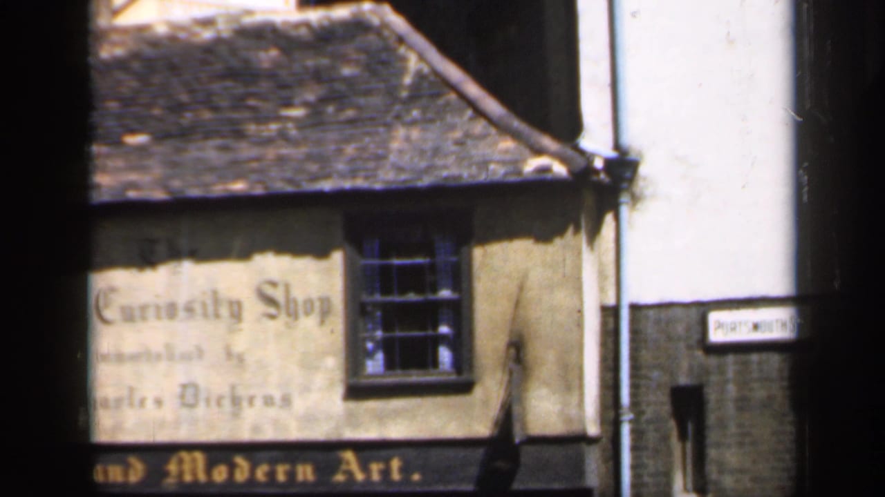 Shopfront in England with faded Old Curiosity Shop lettering, a dark window, tiled roof, and an adjacent street sign visible at frame edge.
