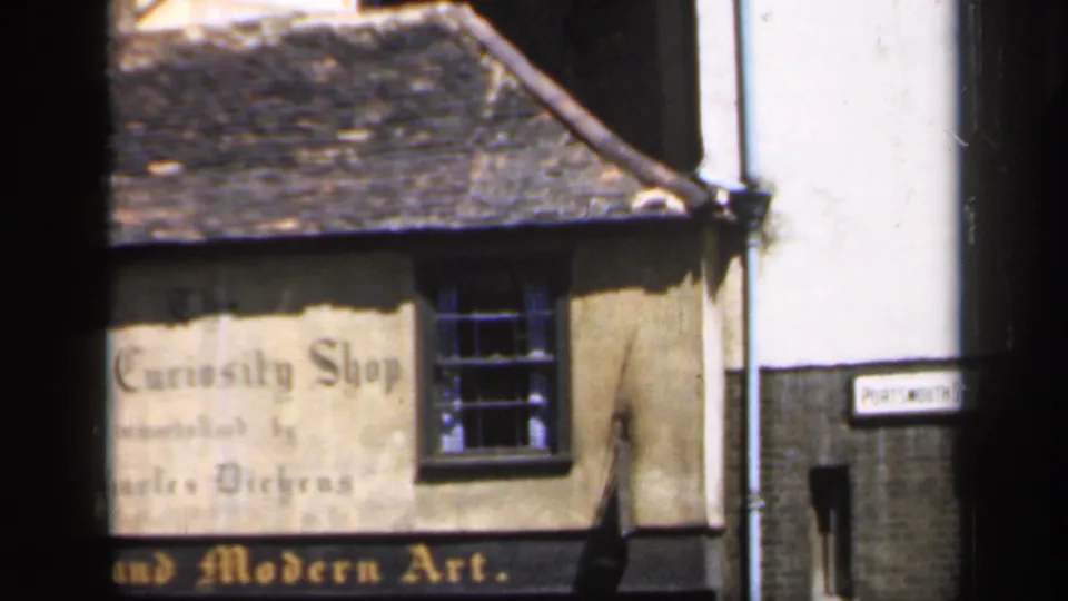 Shopfront in England with faded Old Curiosity Shop lettering, a dark window, tiled roof, and an adjacent street sign visible at frame edge.