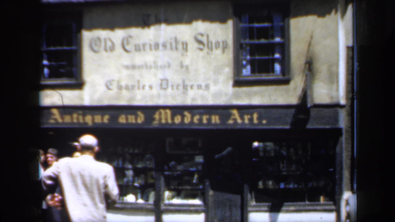 Frame shows The Old Curiosity Shop exterior in England, with Antique and Modern Art lettering, display windows, and adults standing or walking nearby.