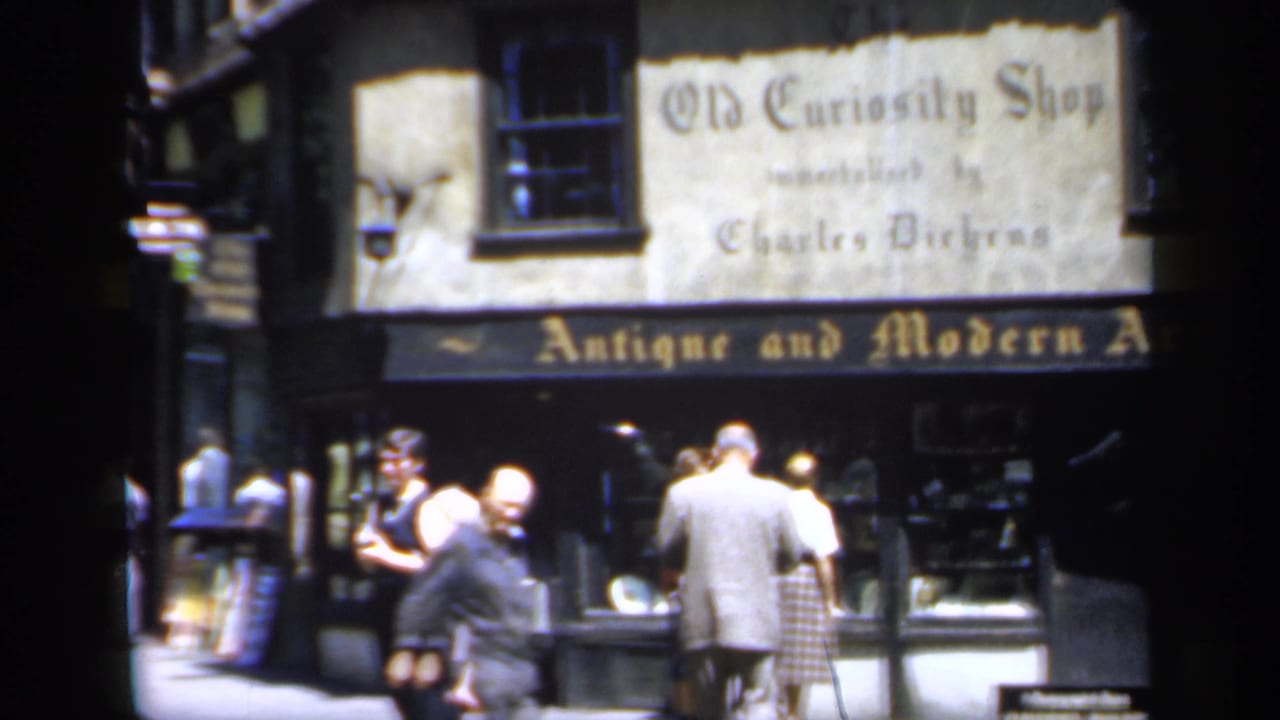1949 England home-movie frame with several pedestrians moving past the Old Curiosity Shop and its antique art storefront signage.