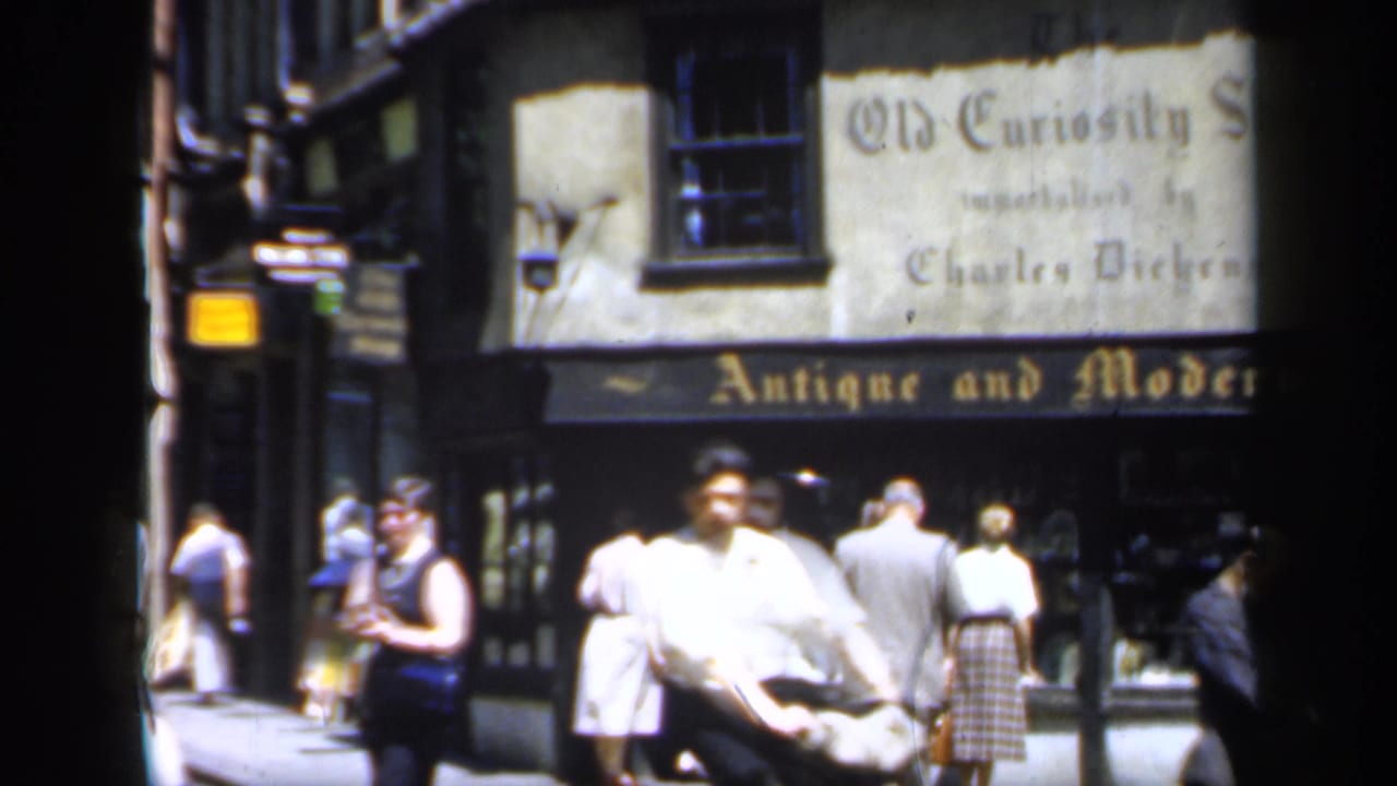 Late-1940s England street scene with a man crossing close to camera and pedestrians outside the Old Curiosity Shop storefront.