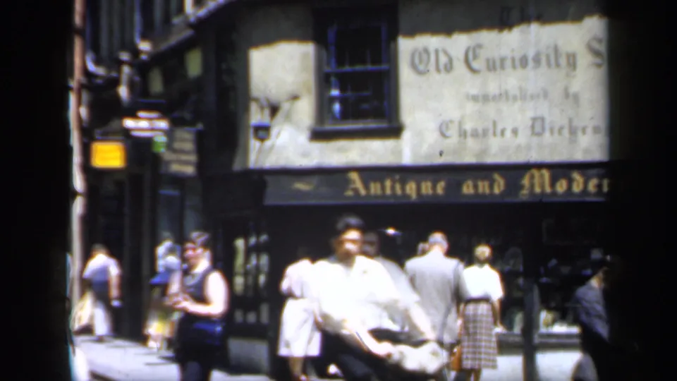 Late-1940s England street scene with a man crossing close to camera and pedestrians outside the Old Curiosity Shop storefront.