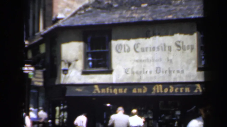 1949 England home-movie frame showing The Old Curiosity Shop facade, antique art signage, and pedestrians in front of the shop.