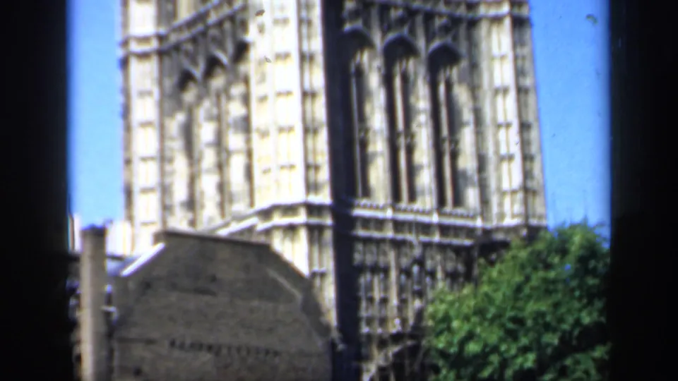 1949 England home-movie frame showing pale Gothic cathedral architecture, arched windows, blue sky, and trees.