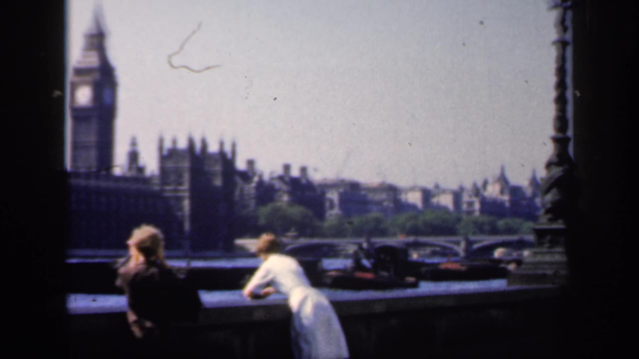 1949 London riverside scene with tourists in the foreground, Big Ben and Parliament behind them, and cars visible near the bridge area.