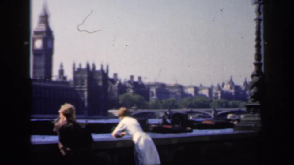 1949 London riverside scene with tourists in the foreground, Big Ben and Parliament behind them, and cars visible near the bridge area.