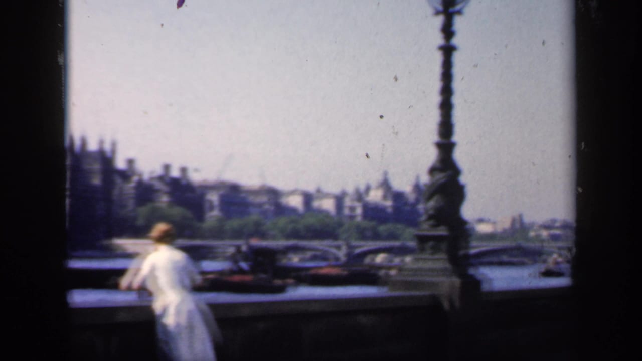 1949 London home-movie frame showing a woman at a Thames bridge railing with ornate lamps, boats, and distant civic buildings.