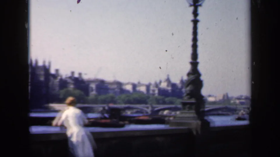 1949 London home-movie frame showing a woman at a Thames bridge railing with ornate lamps, boats, and distant civic buildings.