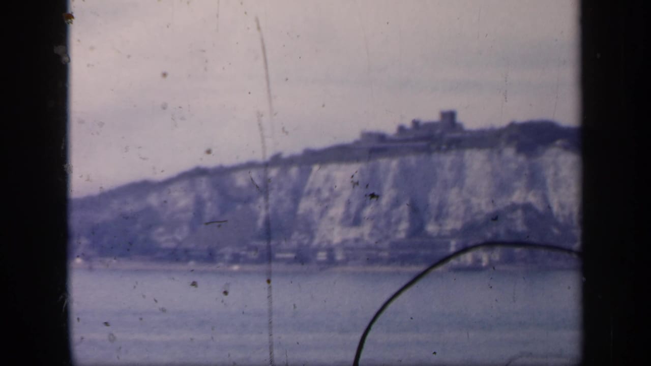 1949 England 8mm frame with chalk cliffs, shoreline buildings, water, and a dark curved foreground mark.