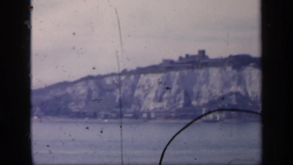 1949 England 8mm frame with chalk cliffs, shoreline buildings, water, and a dark curved foreground mark.