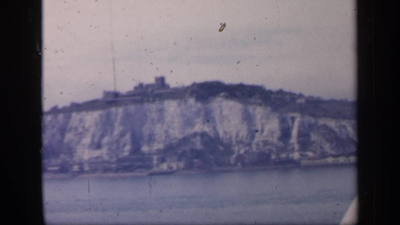 Soft 8mm view of pale coastal cliffs across water with a dark hilltop structure above.