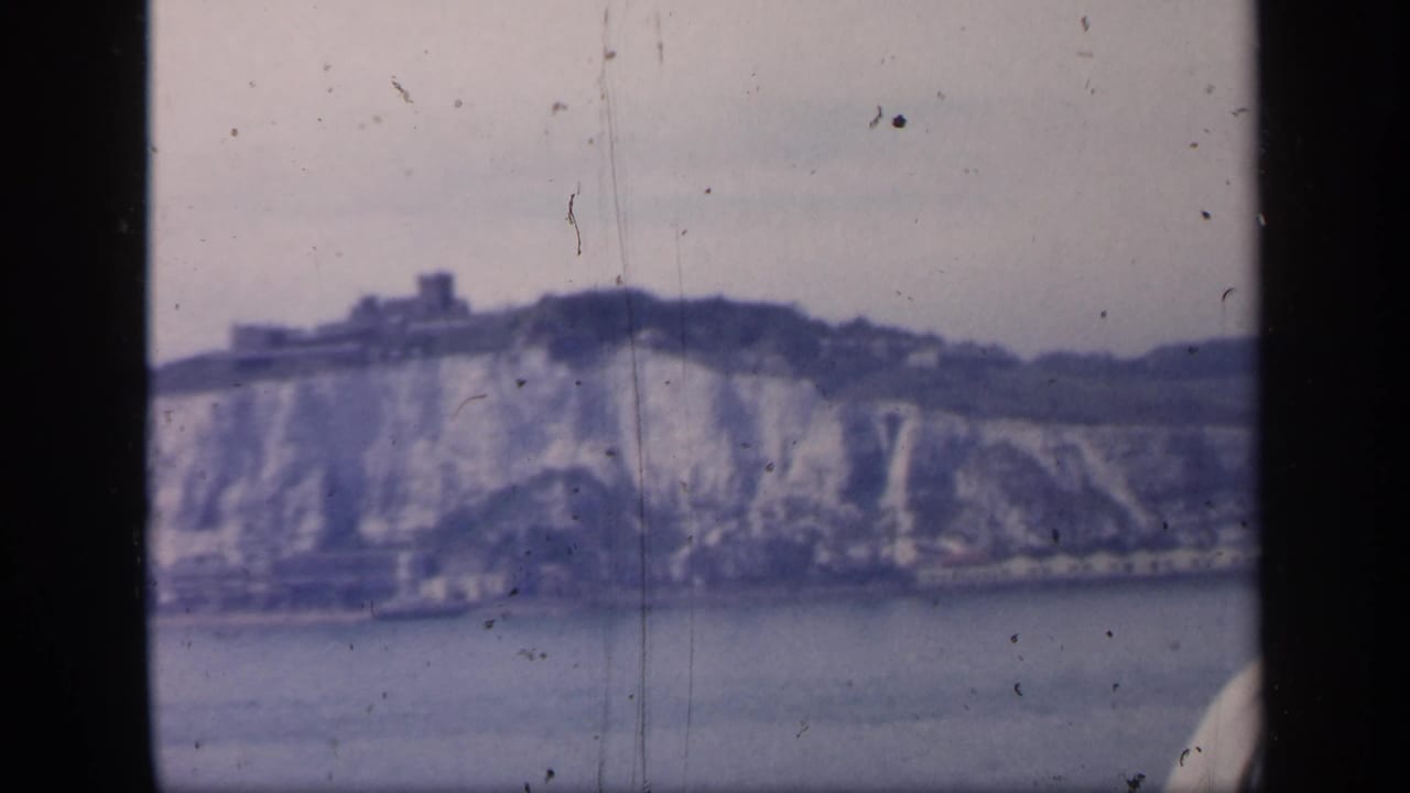 Soft 1949 England travel frame with chalk cliffs, a castle-like building above, and visible scratches from the 8mm source.