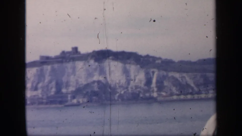 Soft 1949 England travel frame with chalk cliffs, a castle-like building above, and visible scratches from the 8mm source.