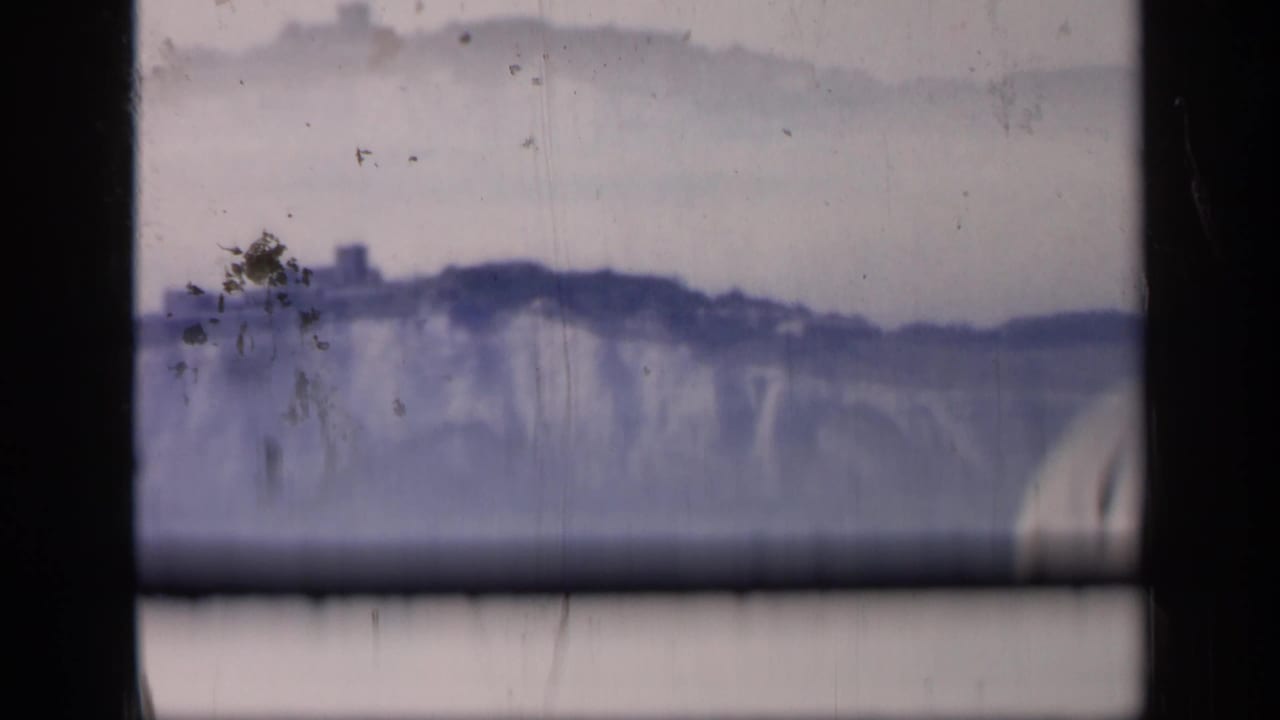 1949 England 8mm frame showing pale coastal cliffs across water, with a dark hilltop structure and heavy film border.