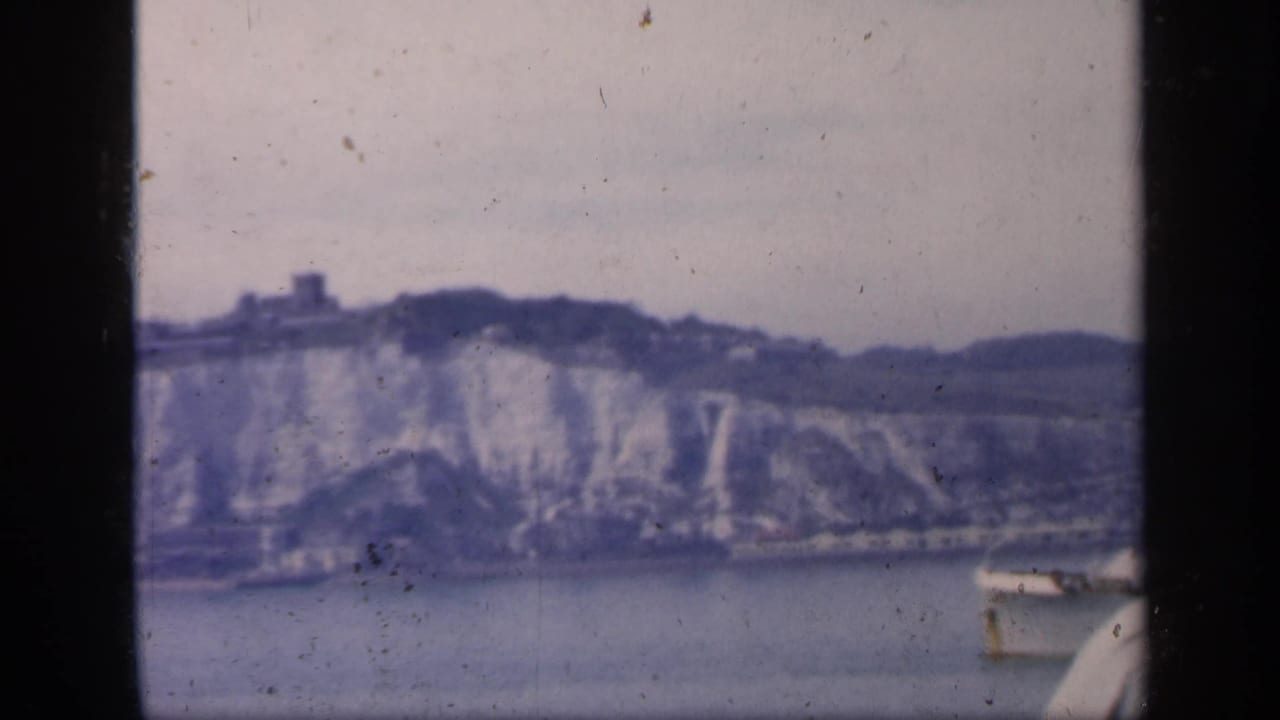 1949 England coastal frame with chalk cliffs in the distance and a small boat entering along the right edge of the water.