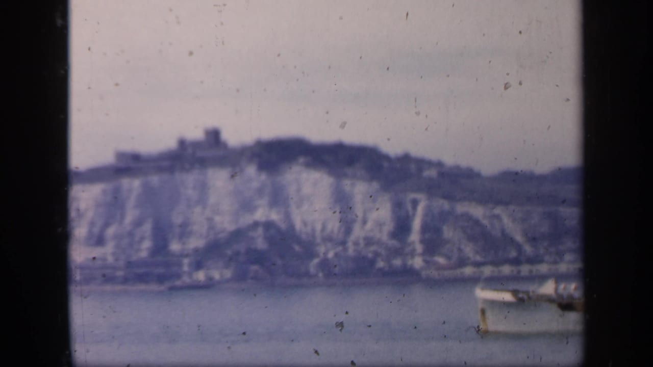 1949 England 8mm frame showing a small boat on calm water beneath pale chalk cliffs and a distant hilltop structure.