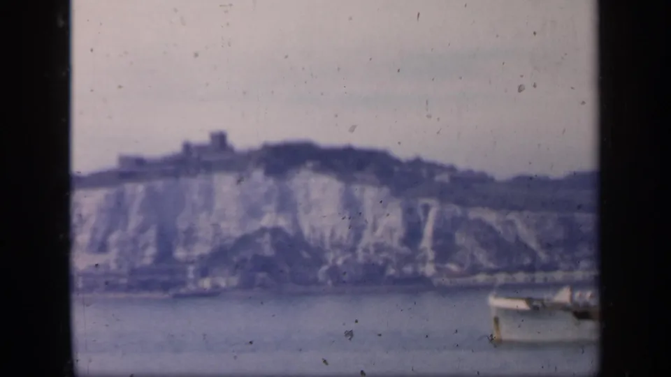 1949 England 8mm frame showing a small boat on calm water beneath pale chalk cliffs and a distant hilltop structure.