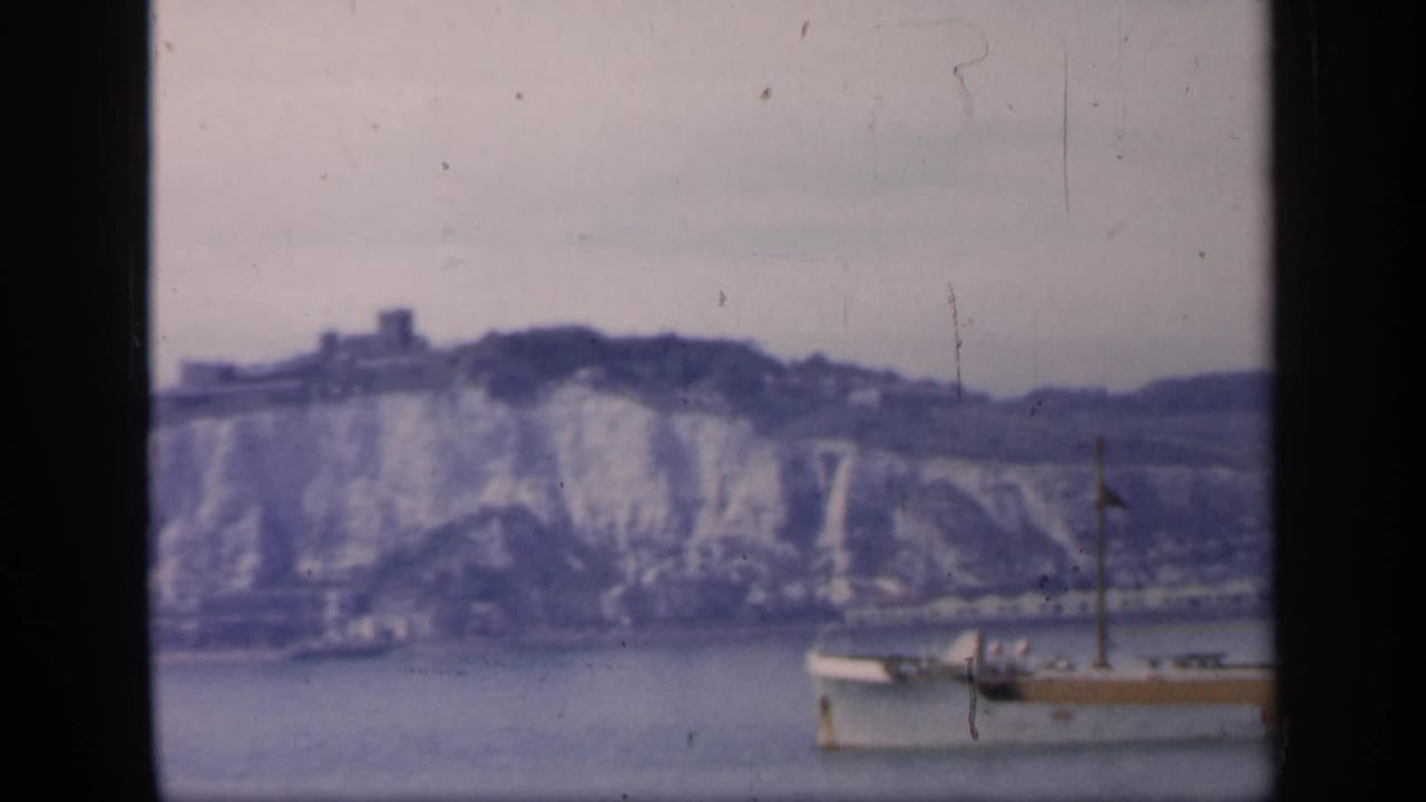 1949 England coastal frame with a small boat in foreground water and white chalk cliffs rising behind it.