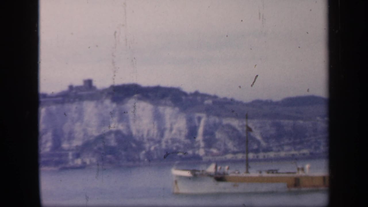 Large ship on water below pale cliffs and a distant hilltop structure.