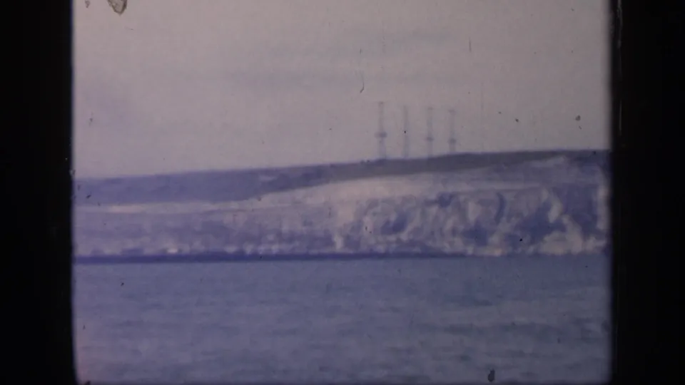 1949 England 8mm frame showing a distant pale cliff beyond the sea, with faint industrial towers or masts along the ridge.