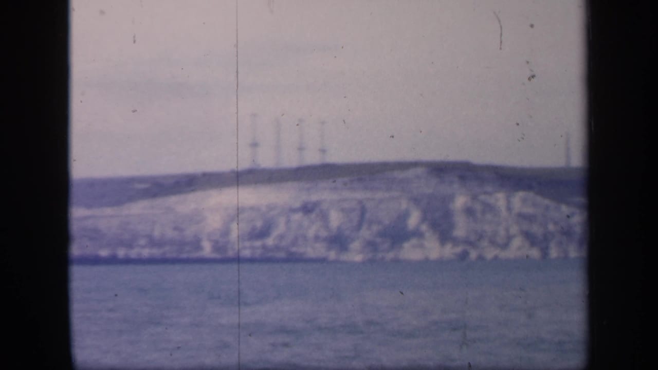 A bluish 1949 England home-movie frame with open water below a pale cliff and several faint towers or masts on the horizon.