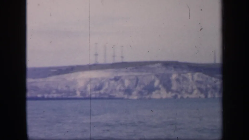 A bluish 1949 England home-movie frame with open water below a pale cliff and several faint towers or masts on the horizon.