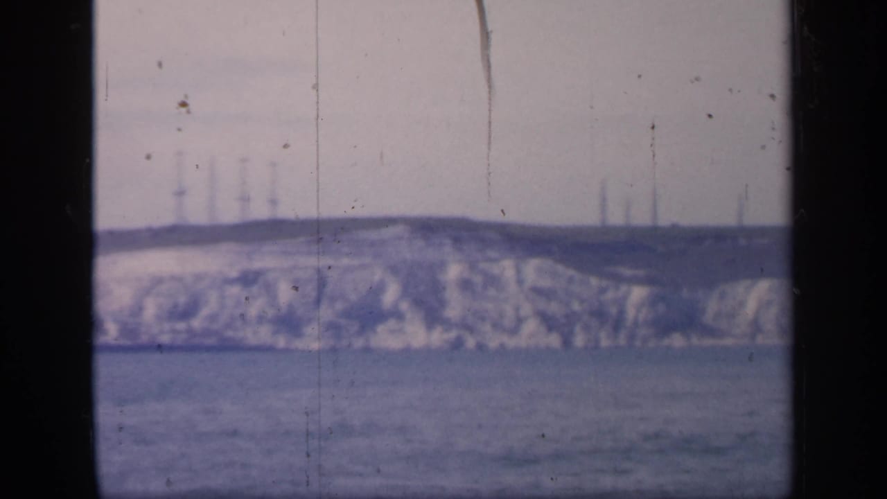 Final 1949 England 8mm frame showing the same distant cliff and sea, with industrial-looking verticals barely visible along the horizon.