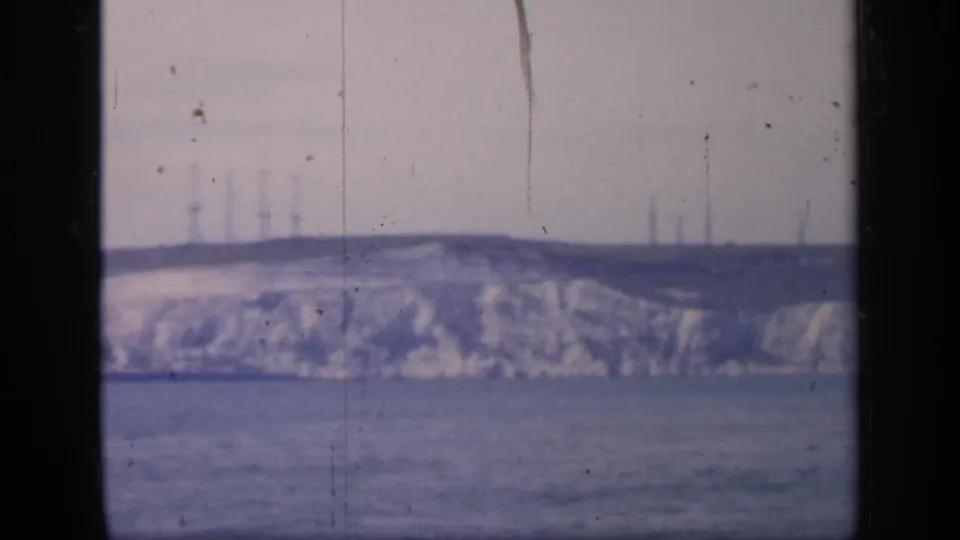 Final 1949 England 8mm frame showing the same distant cliff and sea, with industrial-looking verticals barely visible along the horizon.