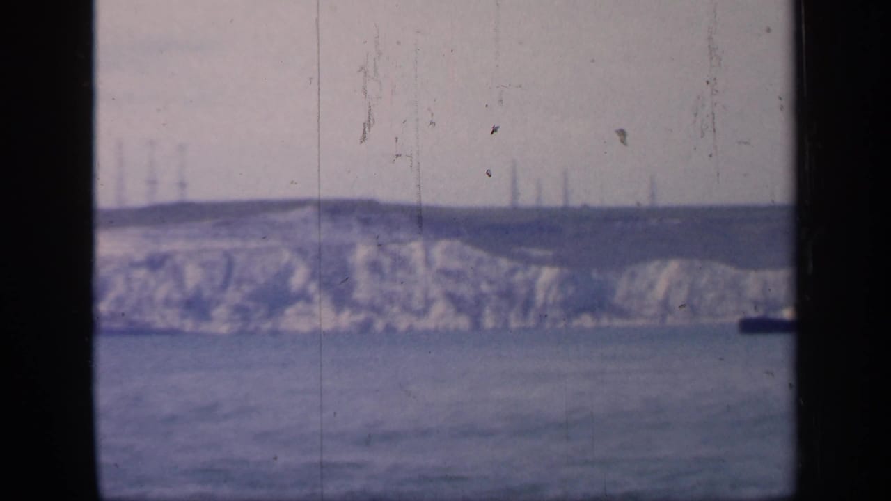 1949 England home-movie frame showing pale chalk cliffs and calm sea viewed through dark window framing, with faint tower structures on the horizon.
