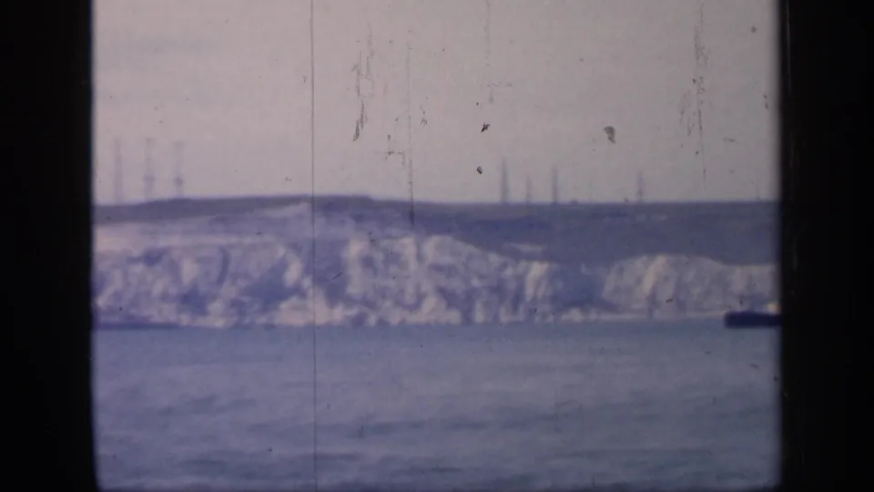 1949 England home-movie frame showing pale chalk cliffs and calm sea viewed through dark window framing, with faint tower structures on the horizon.