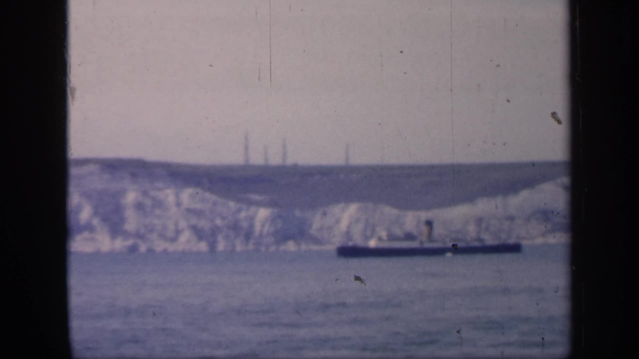 Soft 8mm frame from England in 1949 showing a dark ship beneath chalk cliffs, with tower-like structures faintly visible on the ridge.