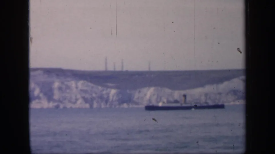 Soft 8mm frame from England in 1949 showing a dark ship beneath chalk cliffs, with tower-like structures faintly visible on the ridge.