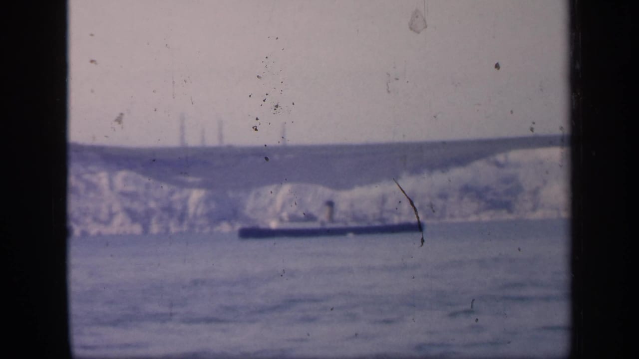 1949 England home-movie frame with a dark ship moving across calm water before chalk cliffs, partially divided by dark window framing.