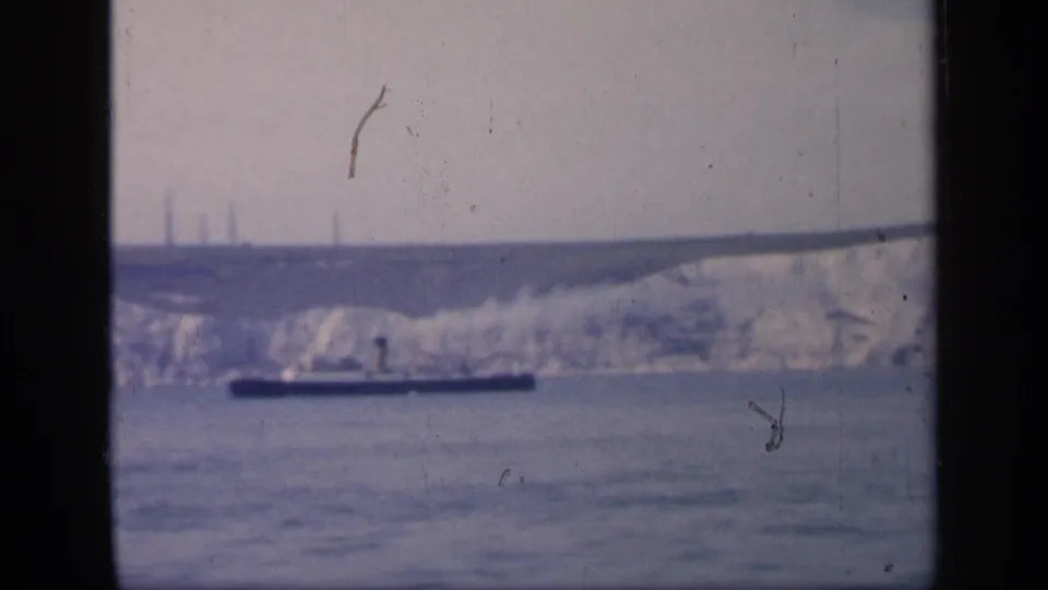 1949 England 8mm frame showing white chalk cliffs beyond open water, with a low dark boat crossing the foreground.