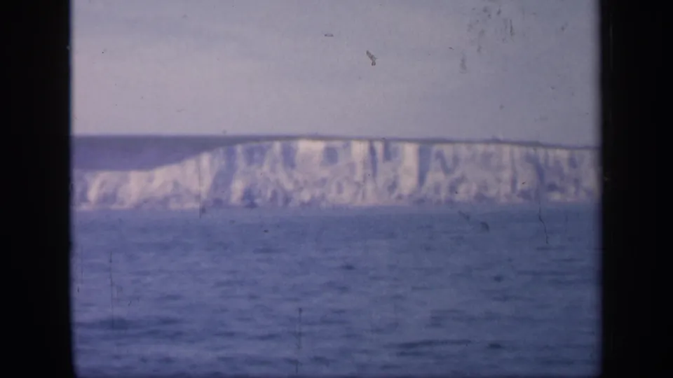 Late-1940s home-movie view of English white cliffs across the water, with a dark vertical vessel window bar in the foreground.