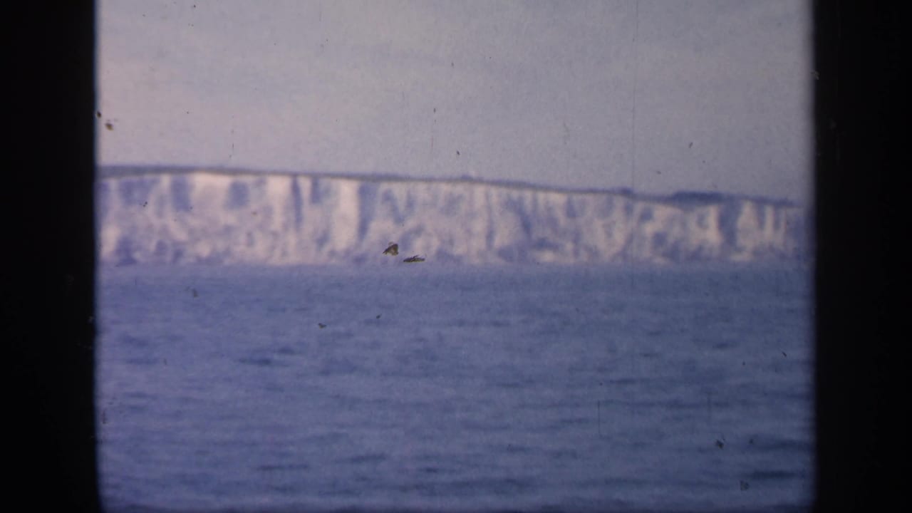 1949 England coastal frame showing white chalk cliffs above calm blue water, with heavy dark borders from the window or film gate.