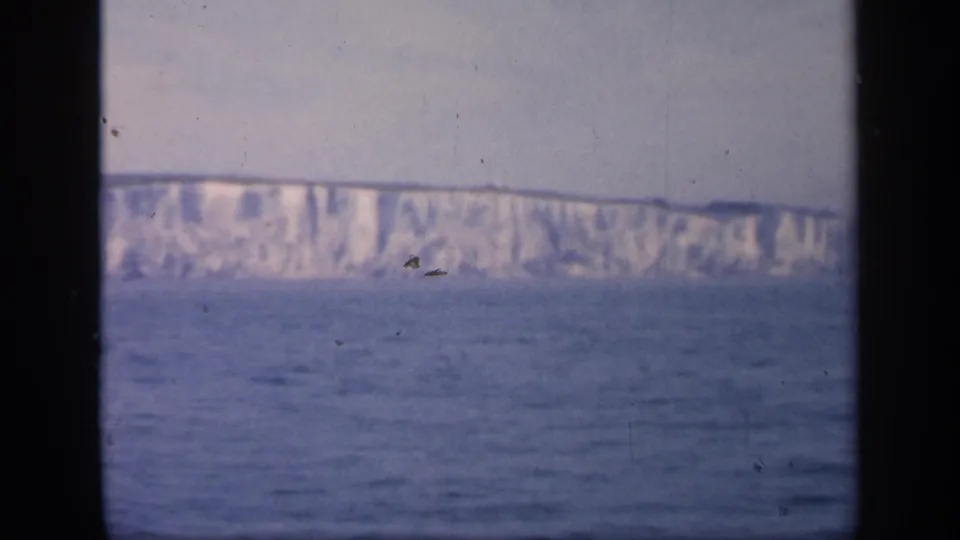1949 England coastal frame showing white chalk cliffs above calm blue water, with heavy dark borders from the window or film gate.
