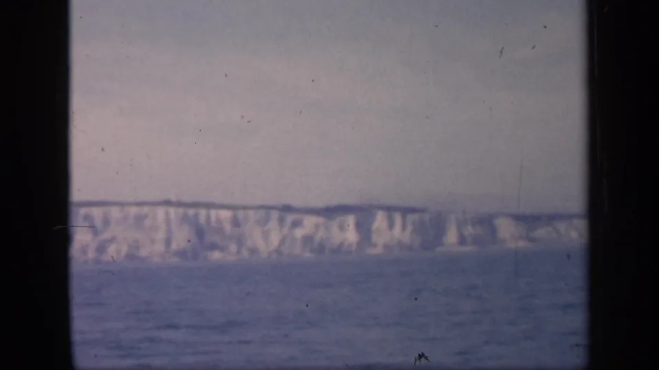 The final sampled frame holds on the English cliff coastline from across the water, with grain, muted color, and gate-edge borders.