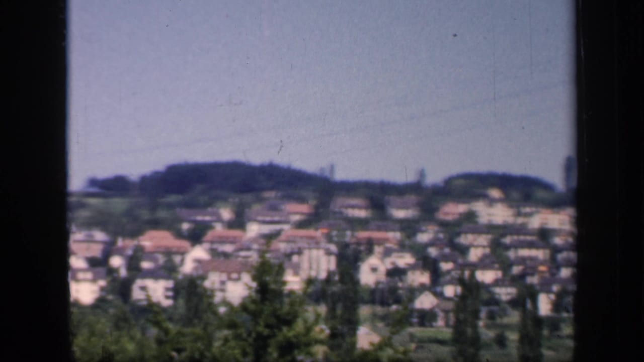 1949 England hillside town view with rows of houses, red roofs, trees, and a distant tower-like shape on the skyline.