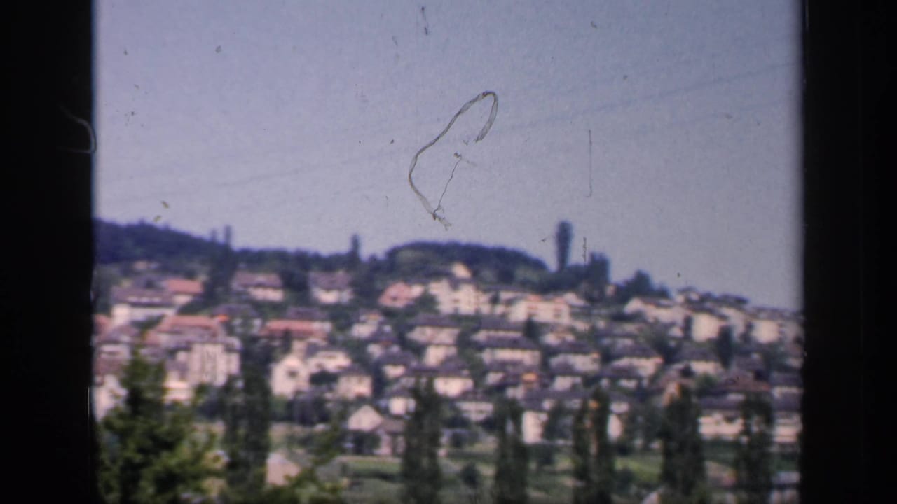 1949 England residential hillside with closely packed homes, foreground trees, and a tall distant structure visible through soft 8mm focus.