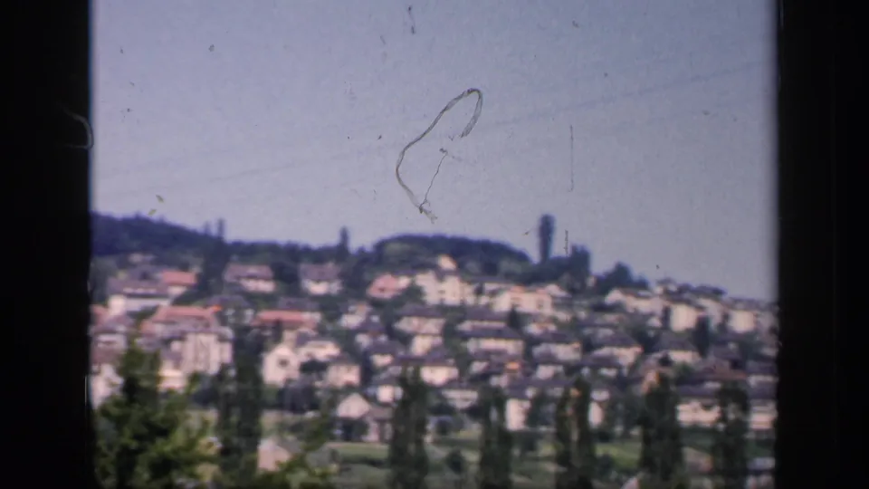 1949 England residential hillside with closely packed homes, foreground trees, and a tall distant structure visible through soft 8mm focus.