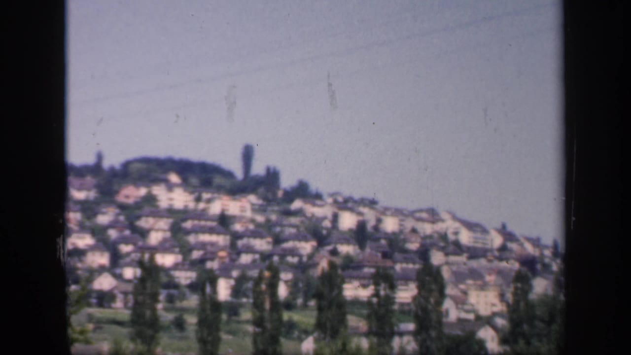 1949 England home-movie frame showing a hillside neighborhood of light-colored houses, trees, rooflines, and open pale sky.
