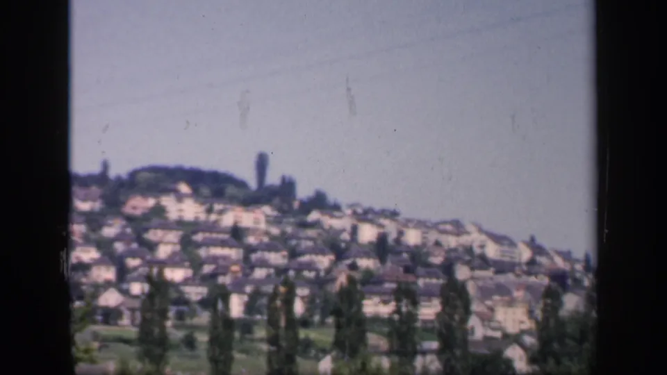 1949 England home-movie frame showing a hillside neighborhood of light-colored houses, trees, rooflines, and open pale sky.