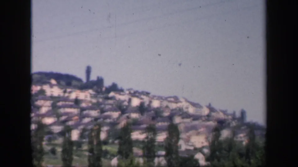1949 England 8mm frame showing a soft distant hillside village, pale houses, tall trees, and clear sky within dark film borders.