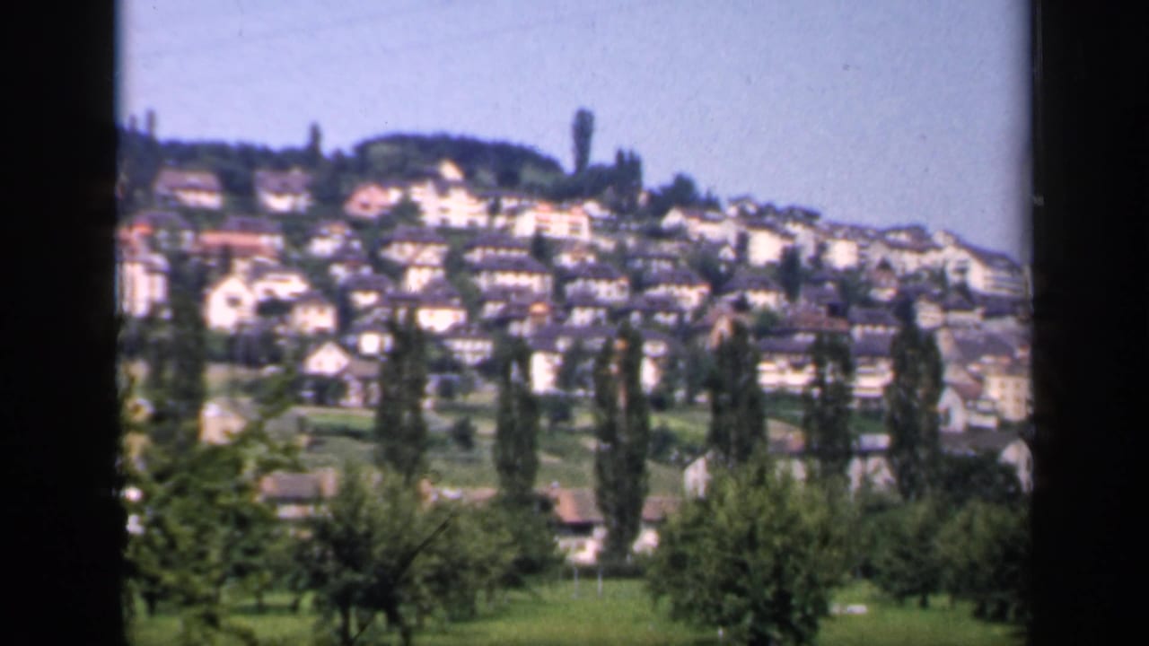1949 England 8mm frame showing a hillside village with dense houses, slender trees, green fields, and a blue sky.
